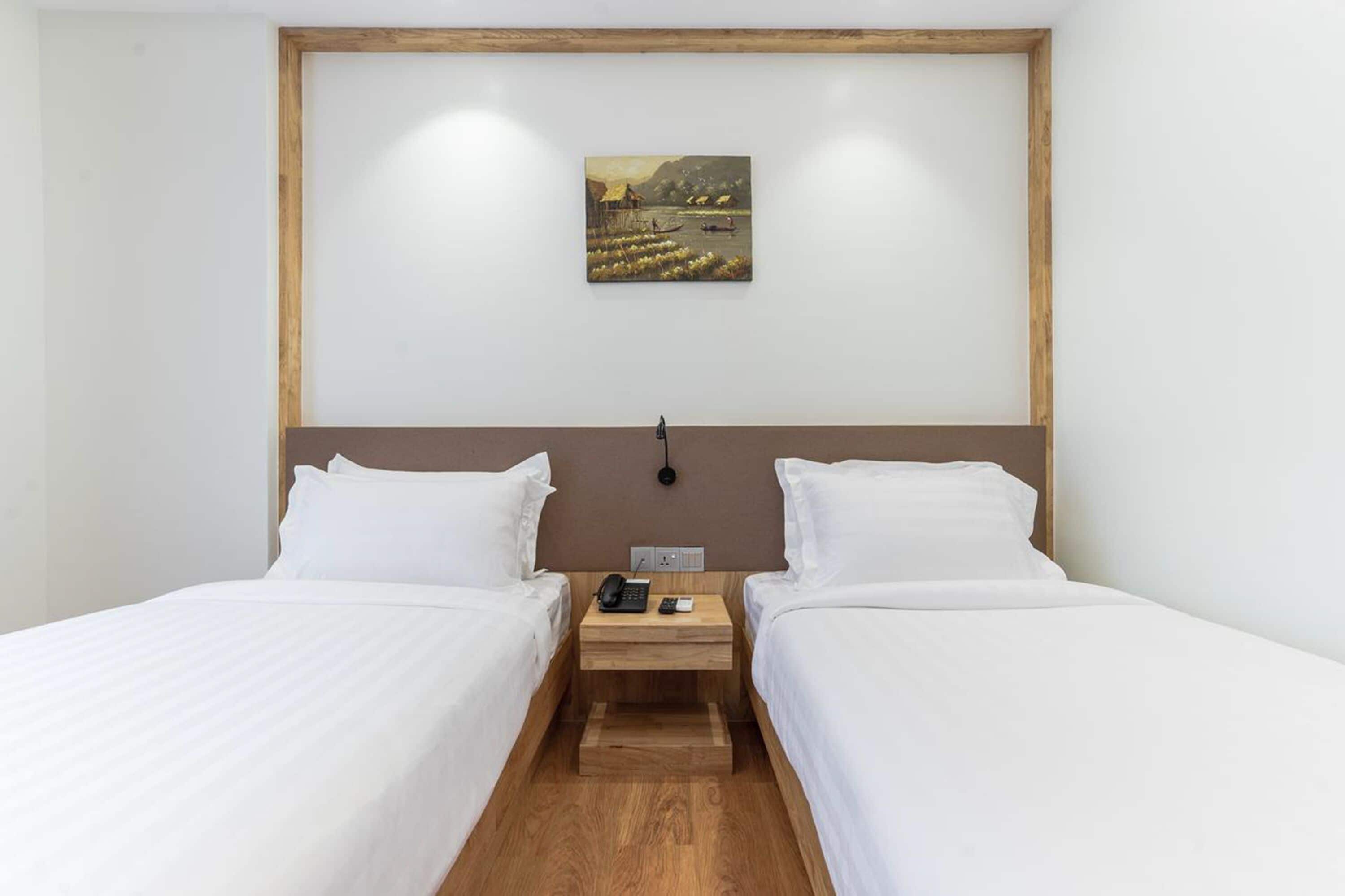 A hotel room with two beds, a wooden headboard, a nightstand with a phone and a lamp, and a painting of a landscape on the wall.
