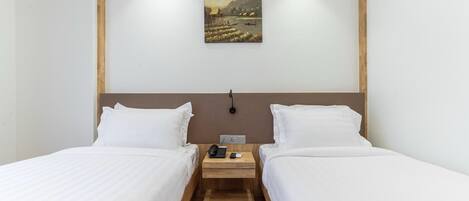 A hotel room with two beds, a wooden headboard, a nightstand with a phone and a lamp, and a painting of a landscape on the wall.