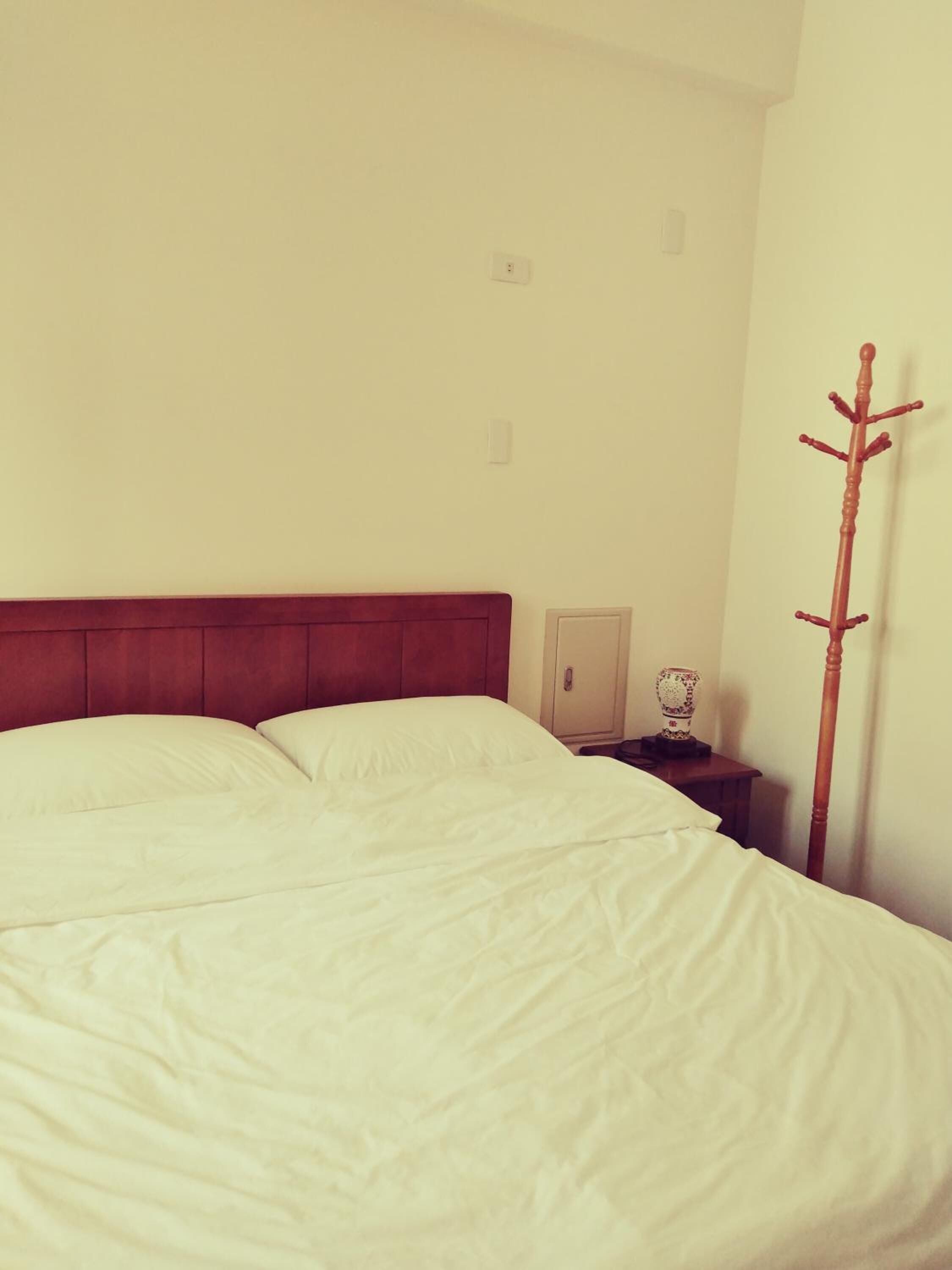 Una cama con sábanas blancas, un cabecero de madera, una mesita de noche con lámpara y un perchero.
