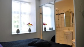 A bedroom with a bed, a window with flowers, a bathroom with a sink and mirror.