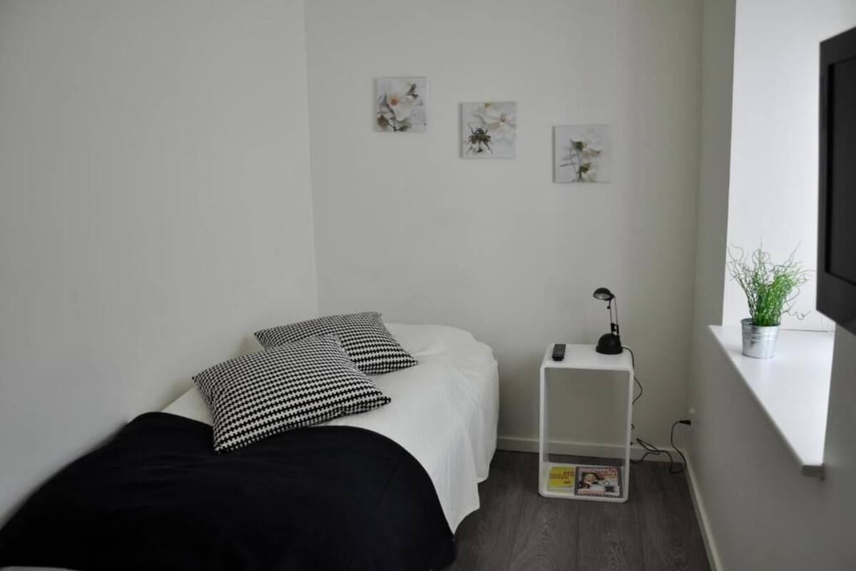 A minimalist bedroom with a bed, black and white pillows, a small white nightstand with a lamp, and wall art.