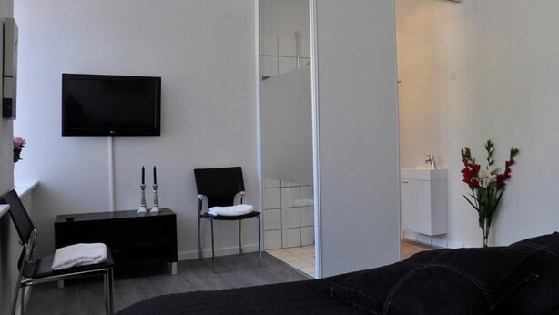 A modern, minimalistic living room with a flat-screen TV, a black sofa, a small black console table, a white chair, and a vase with flowers.