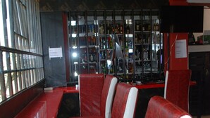A restaurant with red and white chairs, a counter, and a bar area with shelves of bottles. - The REE (Thika)