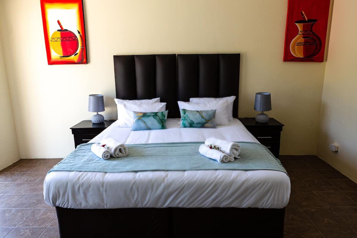 A neatly made bed with white linens and two rolled towels, a black upholstered headboard, and two bedside tables with lamps.