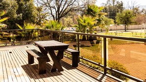 A wooden deck with a picnic table and benches, surrounded by a glass railing and a view of a park. - Avela (Magaliesberg)