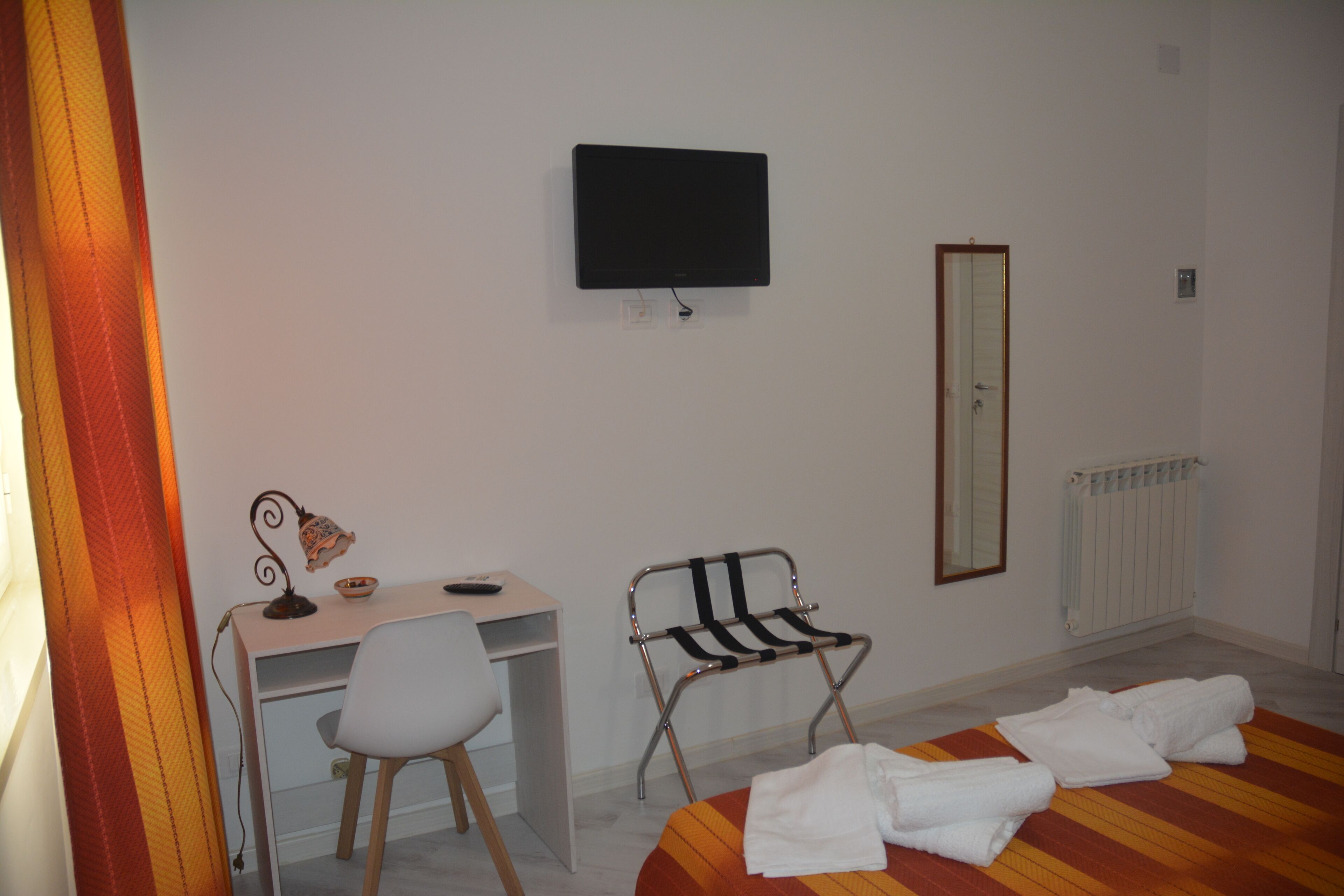 Habitació d'hotel amb un llit, un escriptori amb cadira, una televisió, un mirall i un radiador.