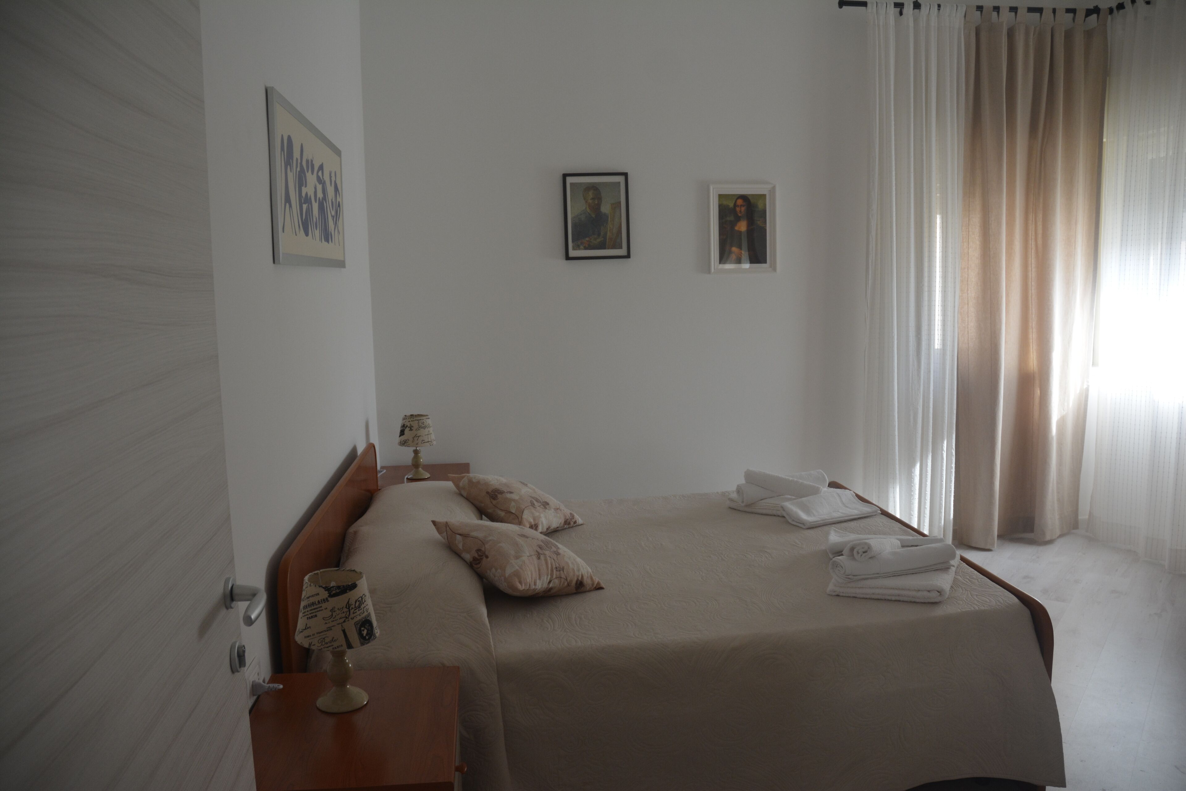 Ein ordentlich bezogenes Bett mit Kissen und gefalteten Handtüchern, ein Nachttisch mit Lampe sowie zwei gerahmte Bilder an der Wand.