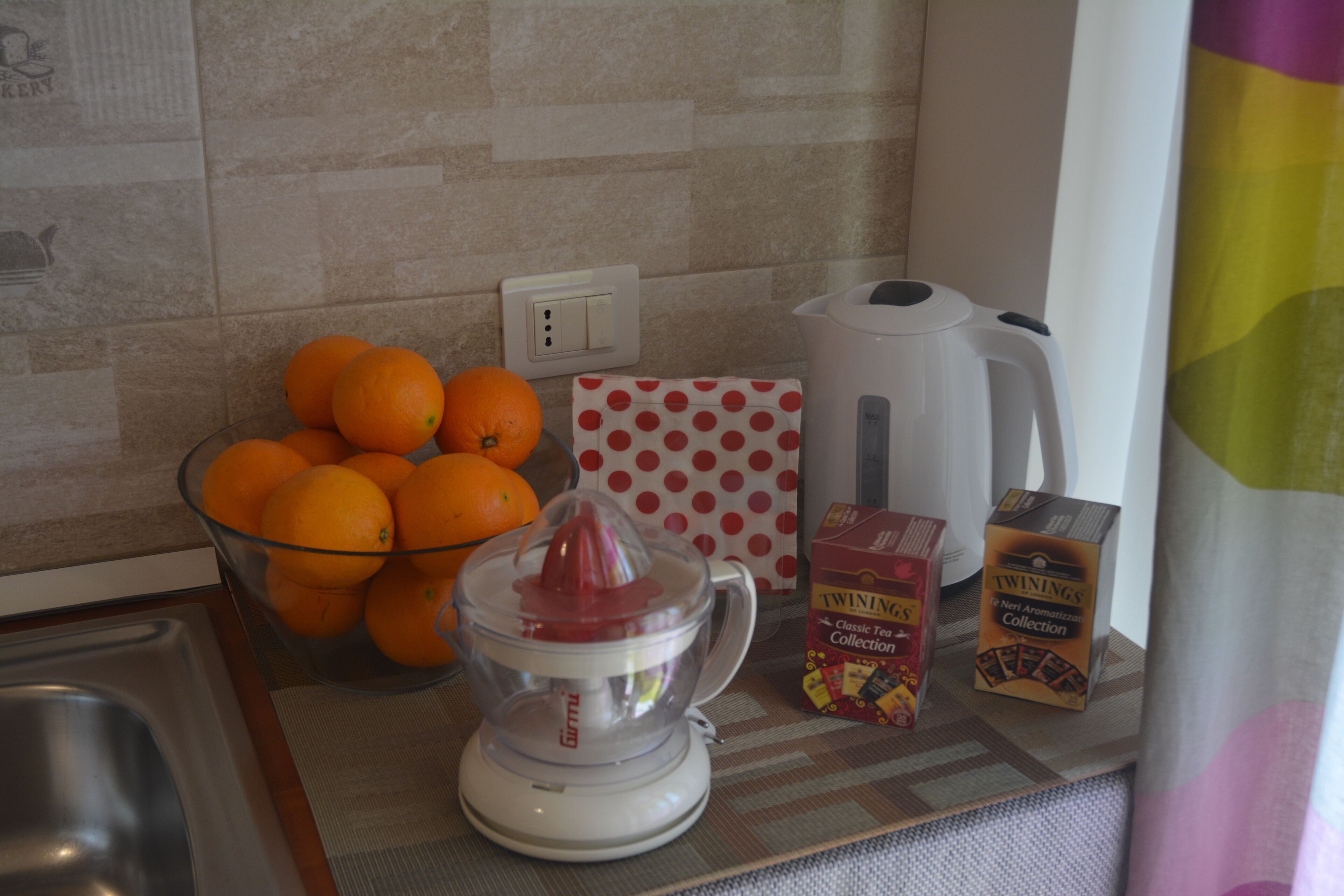 Una encimera de cocina con un tazón de vidrio lleno de naranjas, un exprimidor, una caja de lunares, una tetera y cajas de té Twinings.