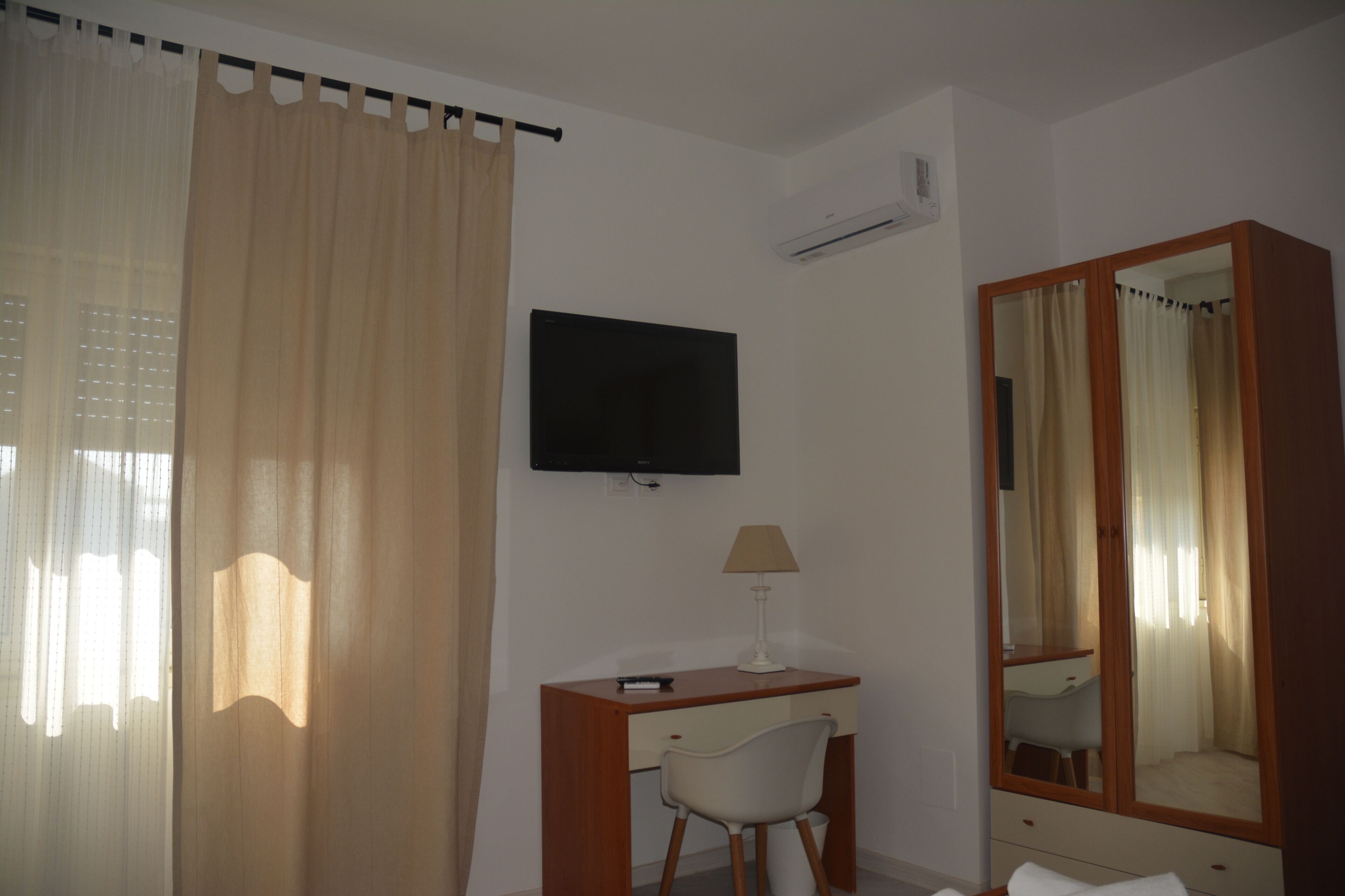 A hotel room with a bed, a desk with a chair, a television, a mirror, and a window with curtains.