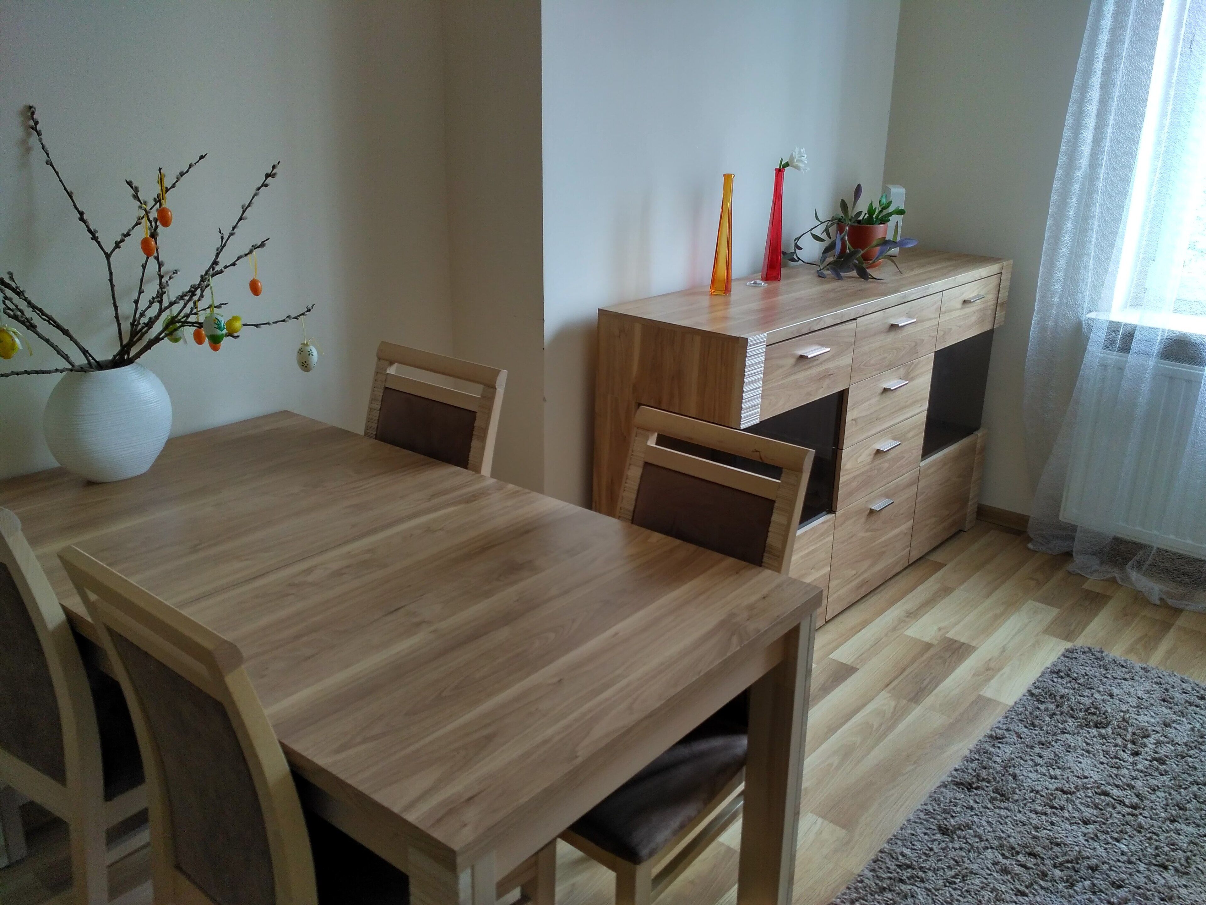 Un comedor con una mesa y sillas de madera, un jarrón blanco con ramas y fruta, y un mueble de madera con cajones.