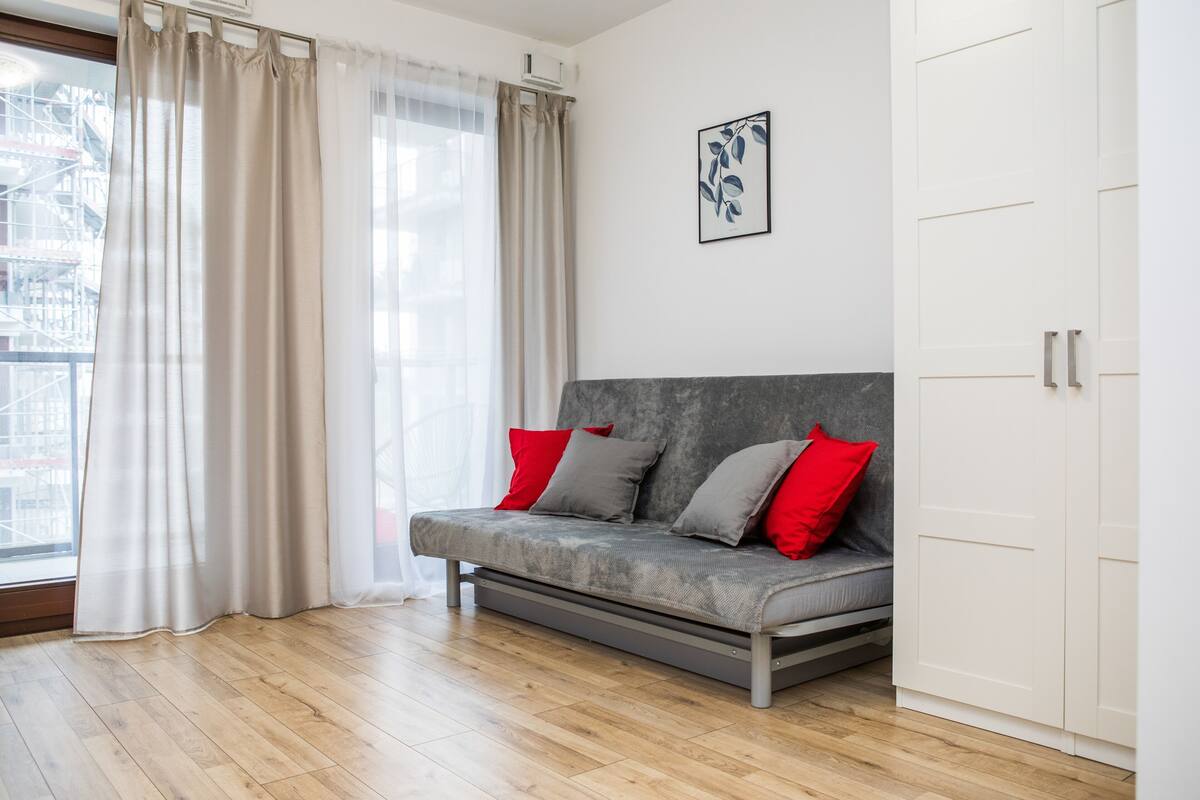 Un salon moderne avec un canapé gris, des coussins rouges et gris, une armoire blanche et une fenêtre ornée de rideaux légers.