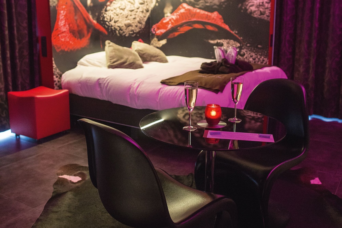 A modern bedroom with a bed, a red ottoman, a black chair, and a table with two glasses of champagne.