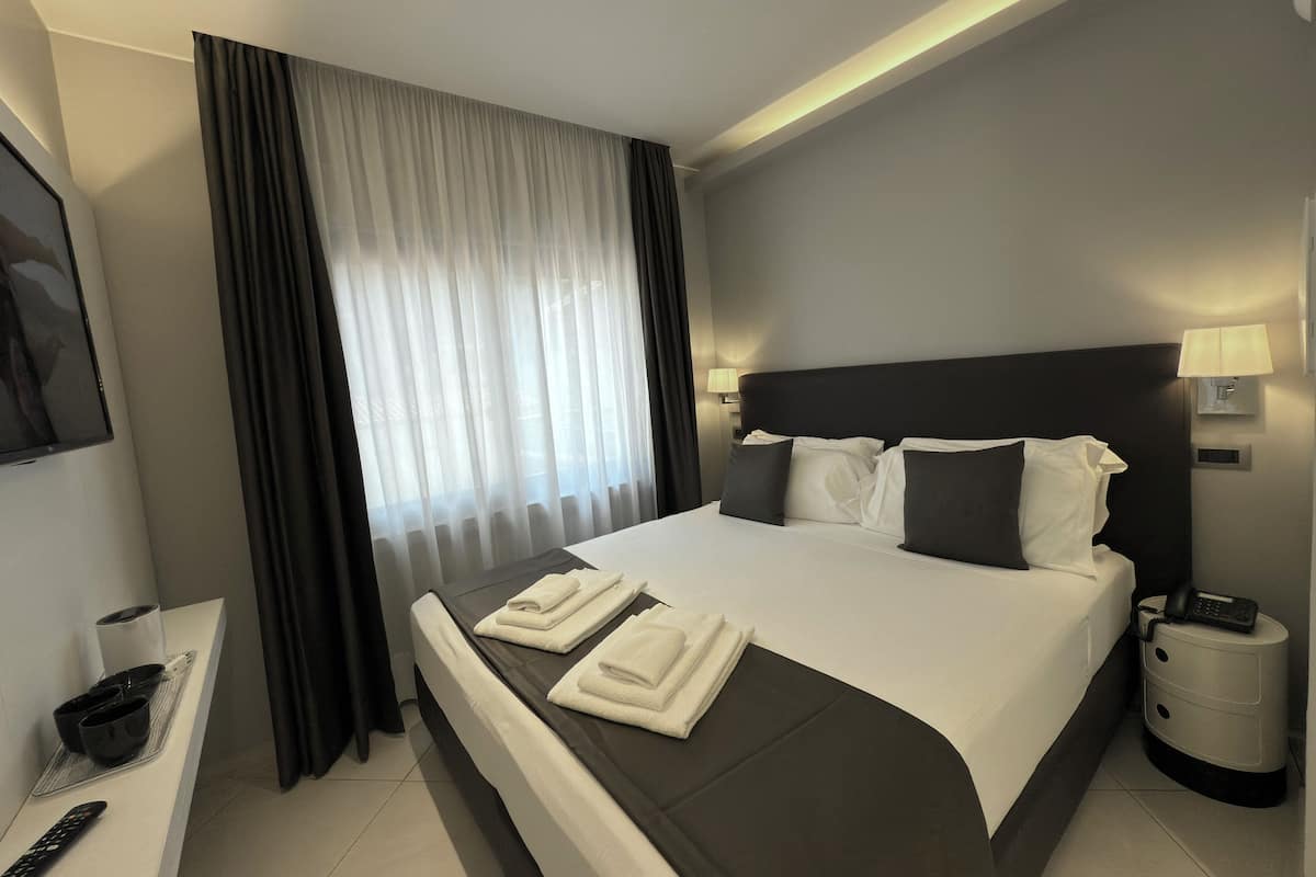 Una habitación de hotel moderna con una cama grande, cabecera oscura, ropa de cama blanca y un televisor de pantalla plana sobre un soporte.