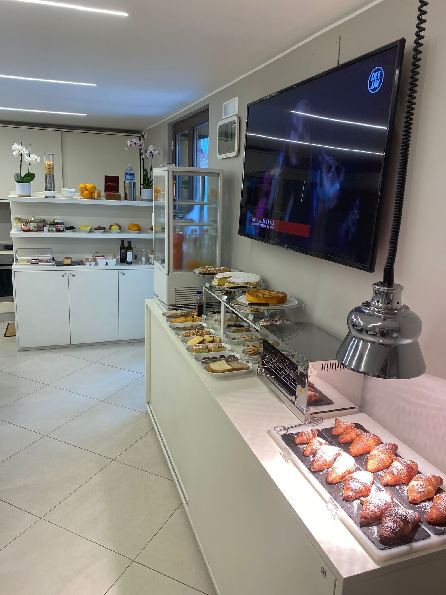Un área de buffet con diversos alimentos, un televisor mostrando un programa y una cocina moderna al fondo.