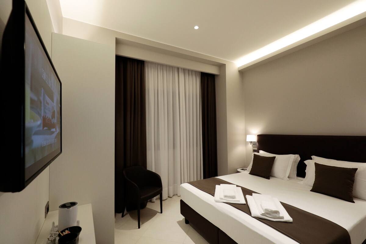 Habitación de hotel con una cama grande, un televisor de pantalla plana montado en la pared, un escritorio con una silla y una ventana con cortinas.