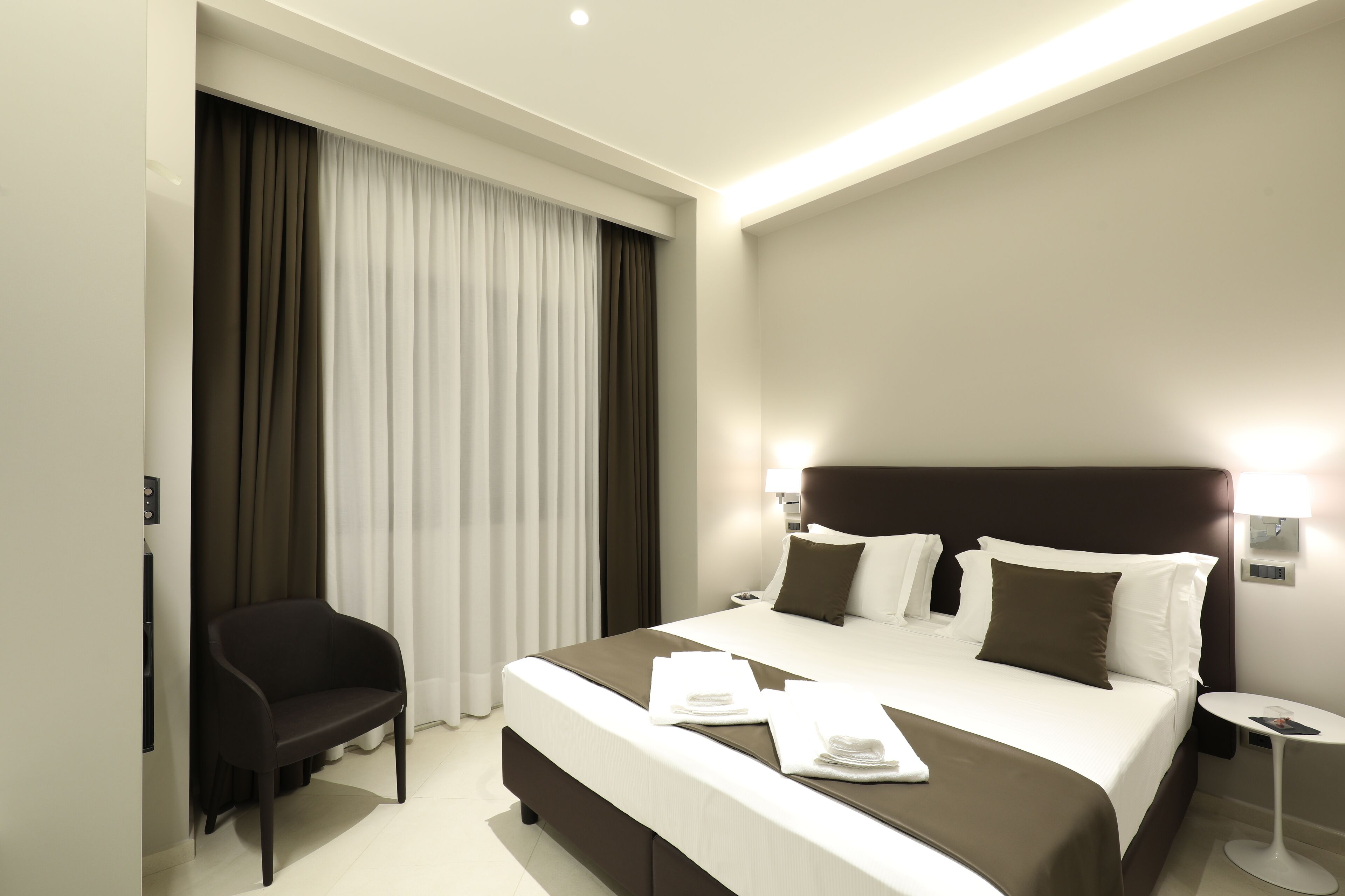 Quarto de hotel moderno com uma cama grande, uma mesinha de cabeceira, uma cadeira e uma janela com cortinas.