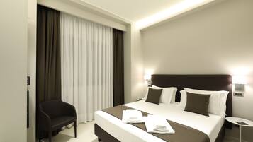 Quarto de hotel moderno com uma cama grande, uma mesinha de cabeceira, uma cadeira e uma janela com cortinas.