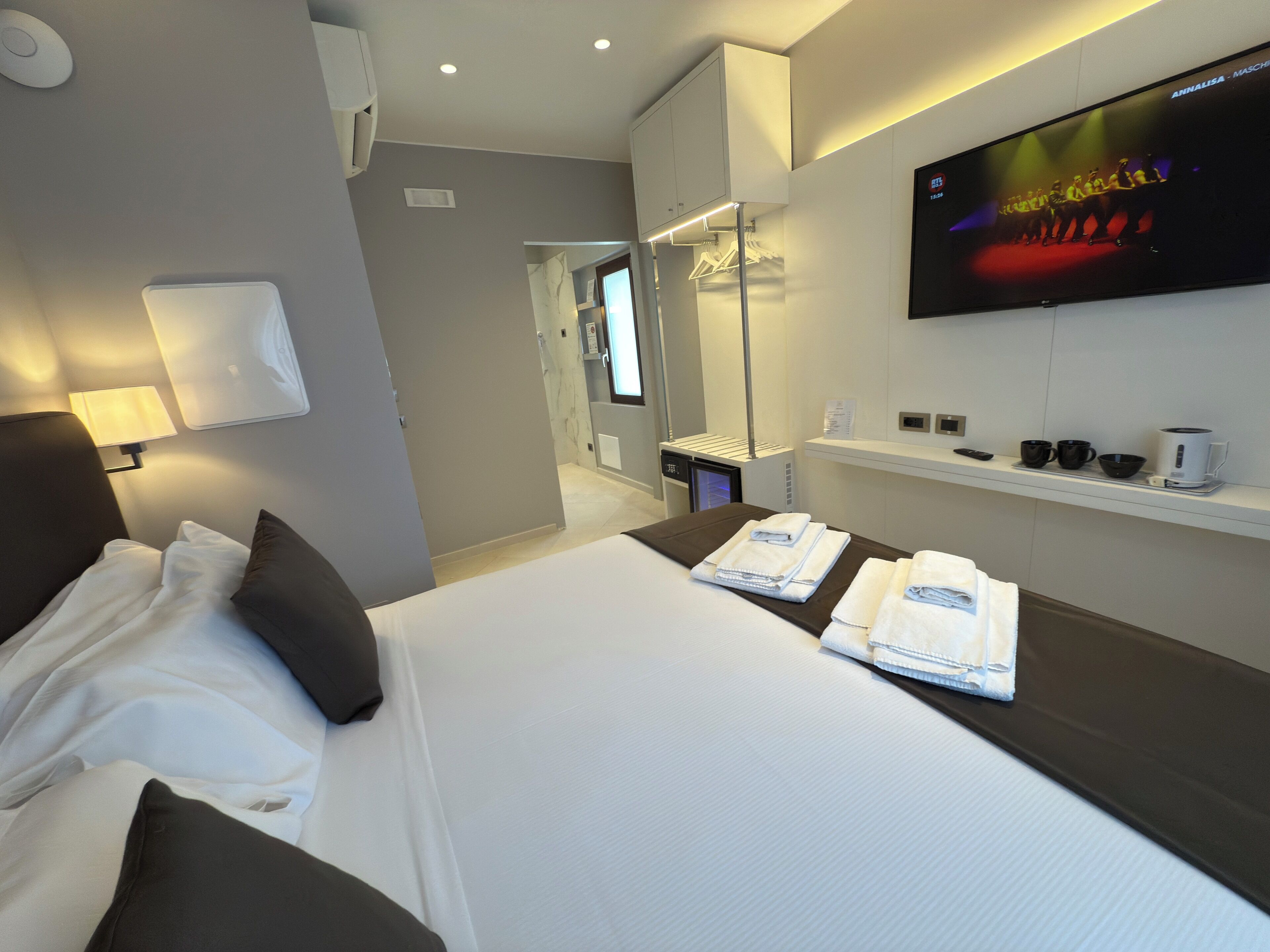 Hotelkamer met een bed, een aan de muur bevestigde televisie, een nachtkastje met handdoeken, een spiegel en een plank met een koffiezetapparaat en kopjes.