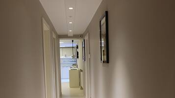 A narrow, well-lit hallway with white walls and a tiled floor, leading to a kitchen area.