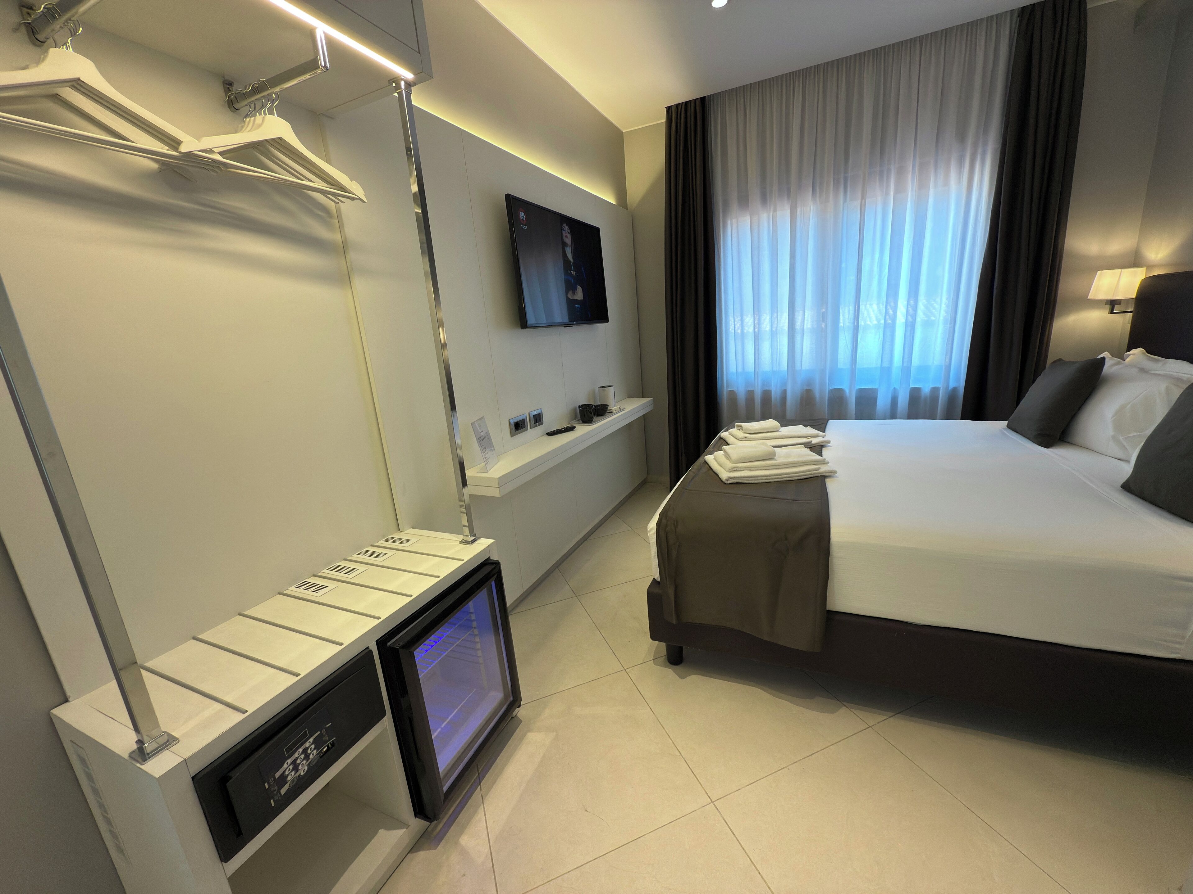 A hotel room with a bed, a TV mounted on the wall, a desk with a computer, and a window with curtains.