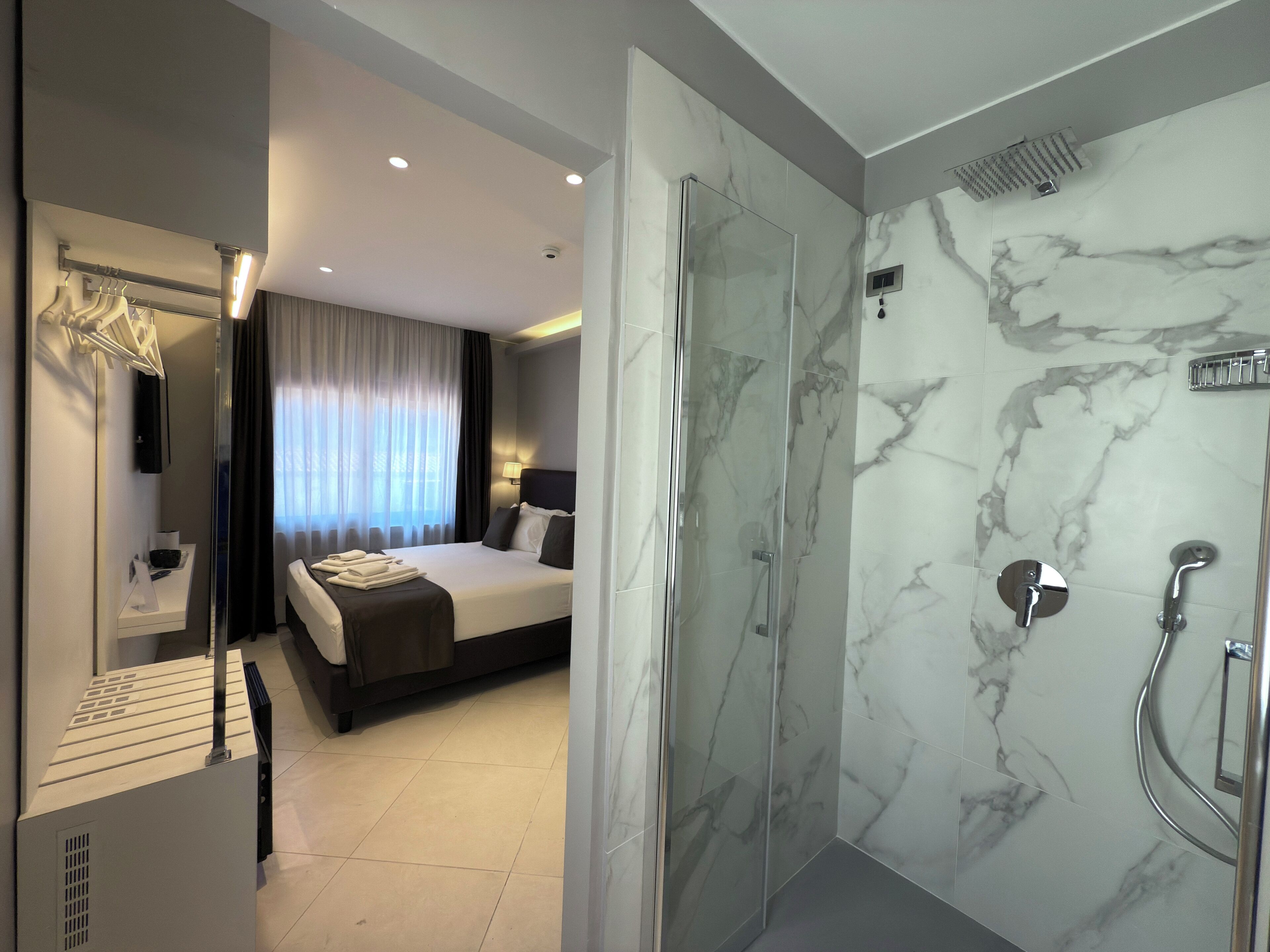 Quarto de hotel moderno com uma cama grande, chuveiro em vidro e paredes de mármore.