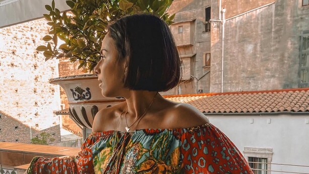 Una persona parada en un balcón con vistas a edificios y una planta en maceta.