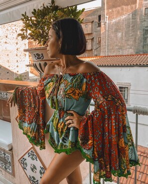 Une personne se tient sur un balcon, d’où l’on peut admirer des immeubles et une plante en pot.