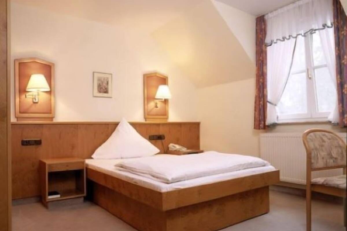 A bedroom with a wooden bed, bedside lamps, a nightstand, a chair, and a window with curtains.