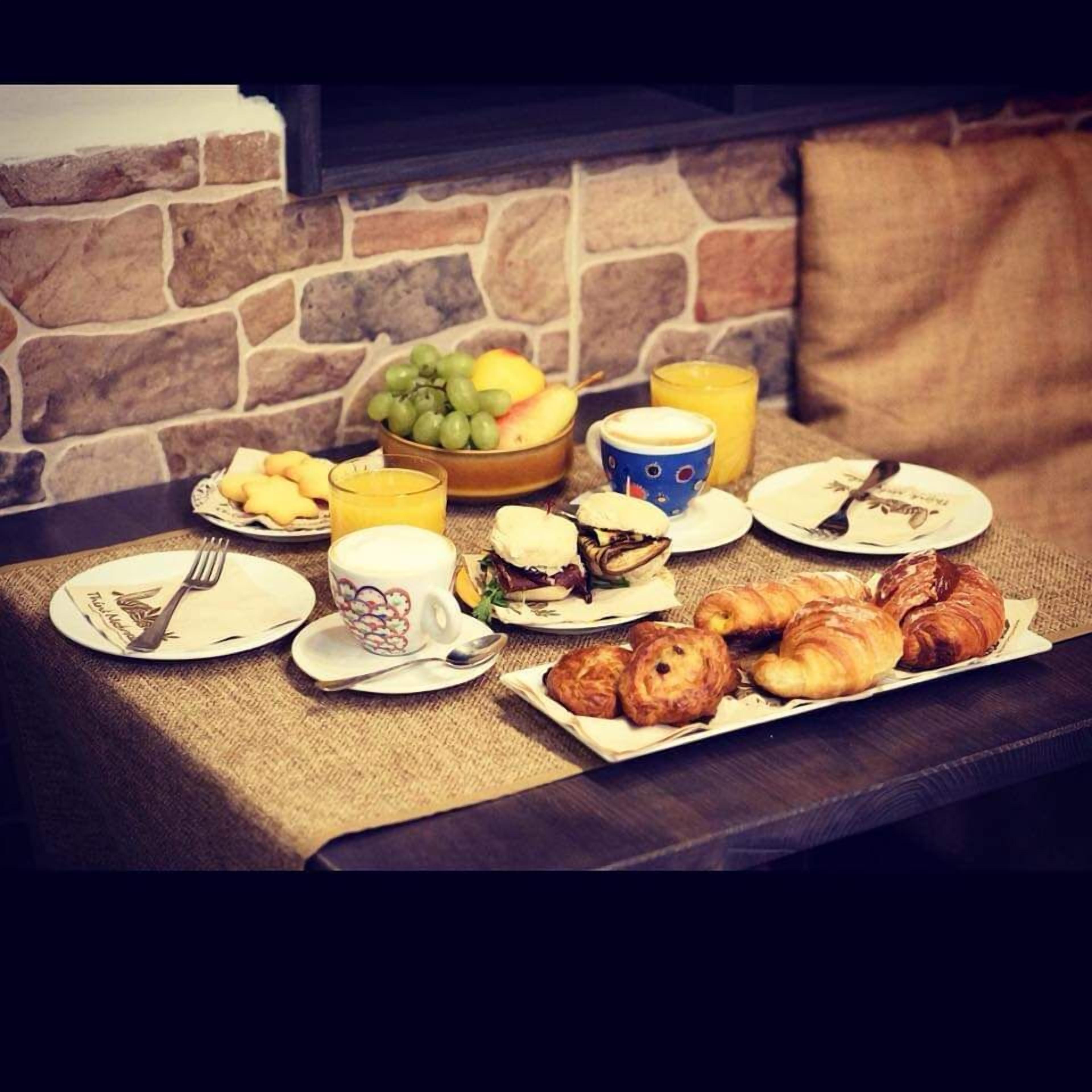 A breakfast setup with croissants, a fruit bowl, a coffee cup, and a juice glass on a table with a stone wall backdrop.