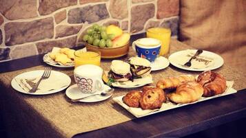 A breakfast setup with croissants, a fruit bowl, a coffee cup, and a juice glass on a table with a stone wall backdrop.