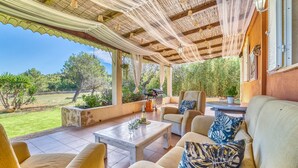 A covered outdoor seating area with a table and chairs, a grill, and a view of the surrounding greenery.