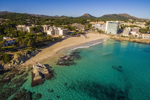 An aerial view of a coastal area with a beach, clear turquoise waters, and buildings in the background.