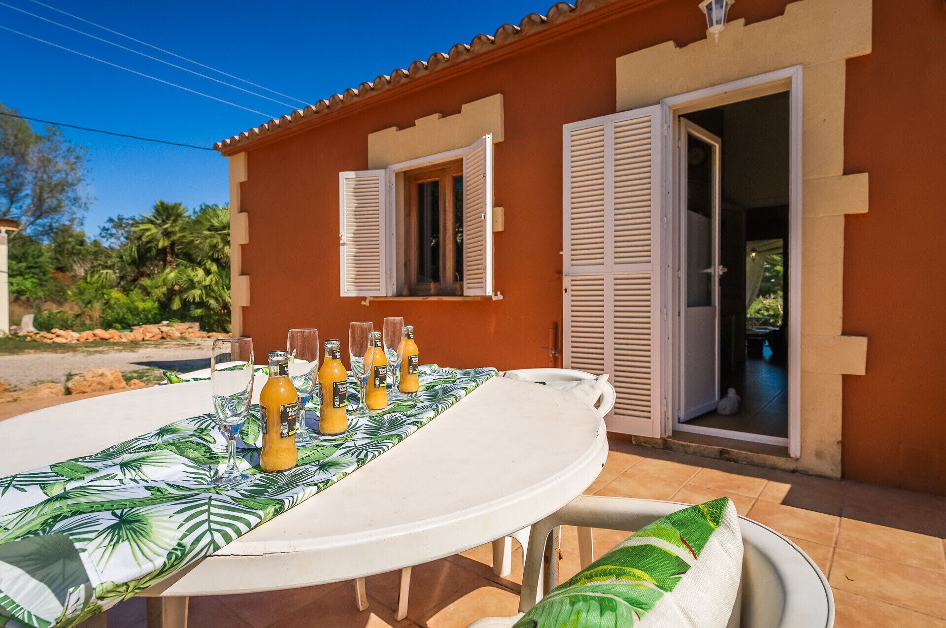 A table set outdoors with a tropical leaf tablecloth, a white chair, and several bottles of orange juice.