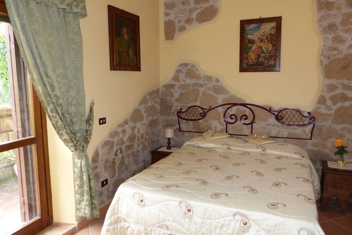 Een slaapkamer met een stenen muur, een bed met een decoratief hoofdeinde, een nachtkastje en twee ingelijste foto’s aan de wand.