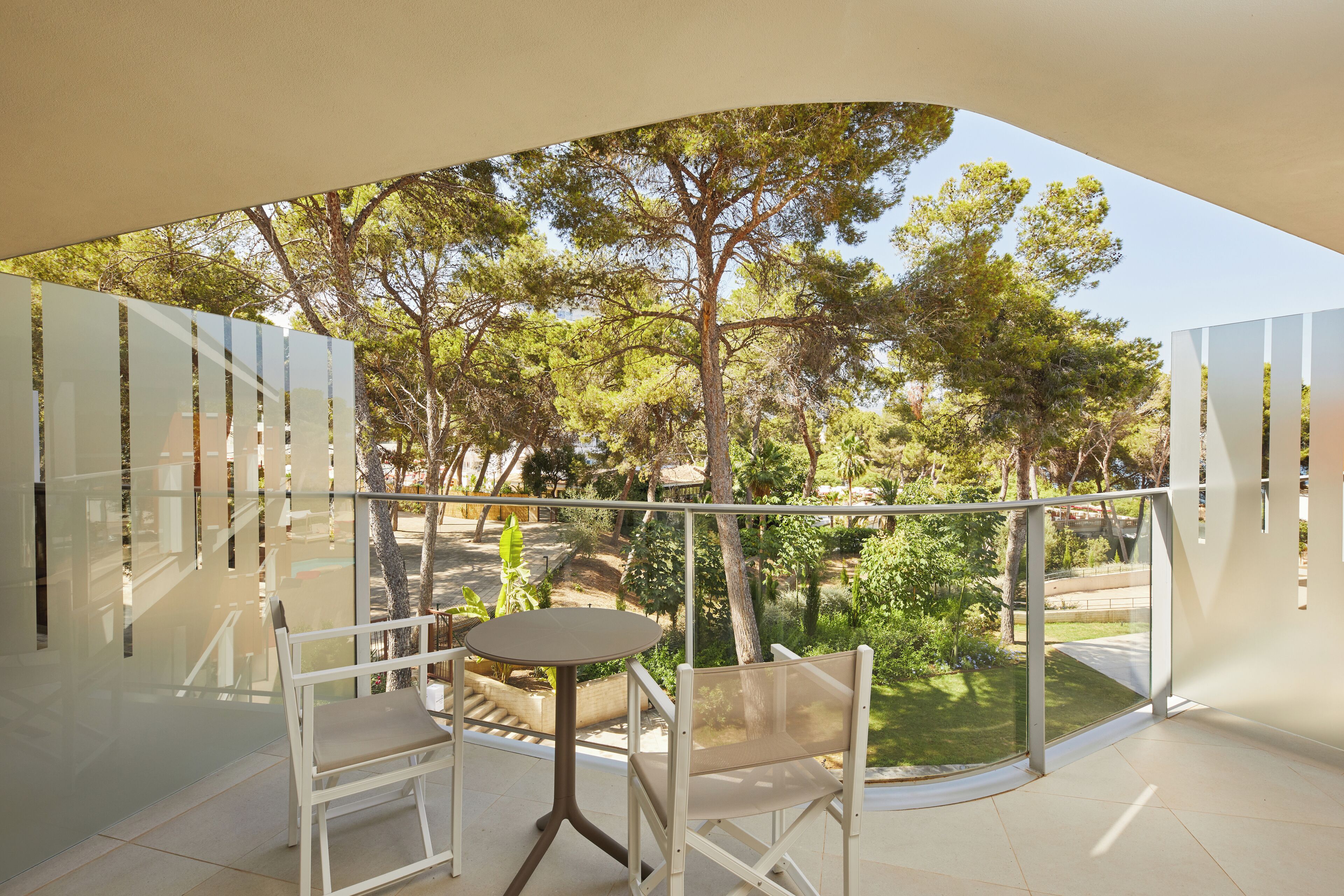 A balcony with a glass railing, a round table, and two chairs, overlooking a garden with trees and a pathway.