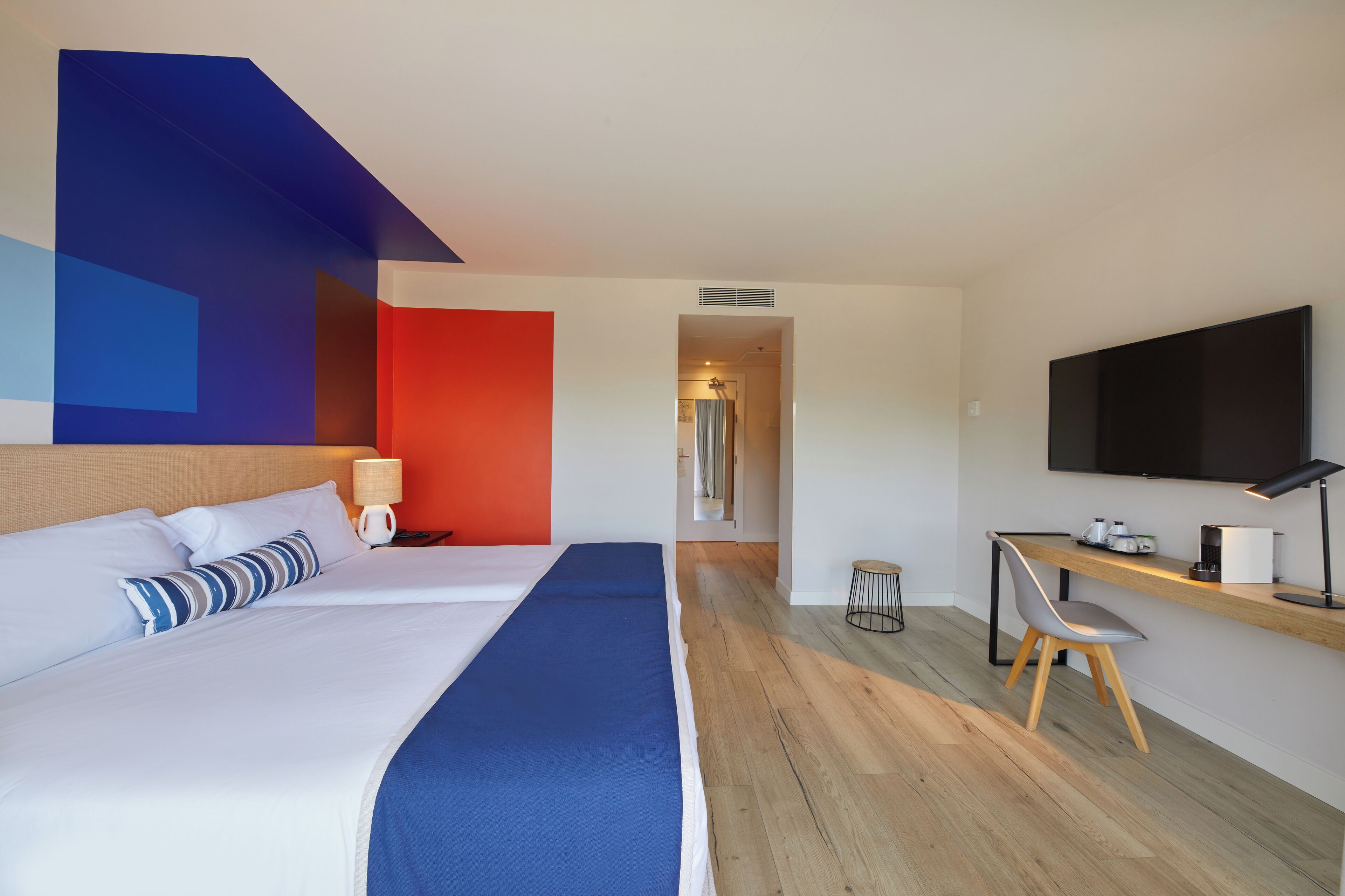 A modern hotel room with a large bed, a desk with a chair, a television, and a view of the hallway.