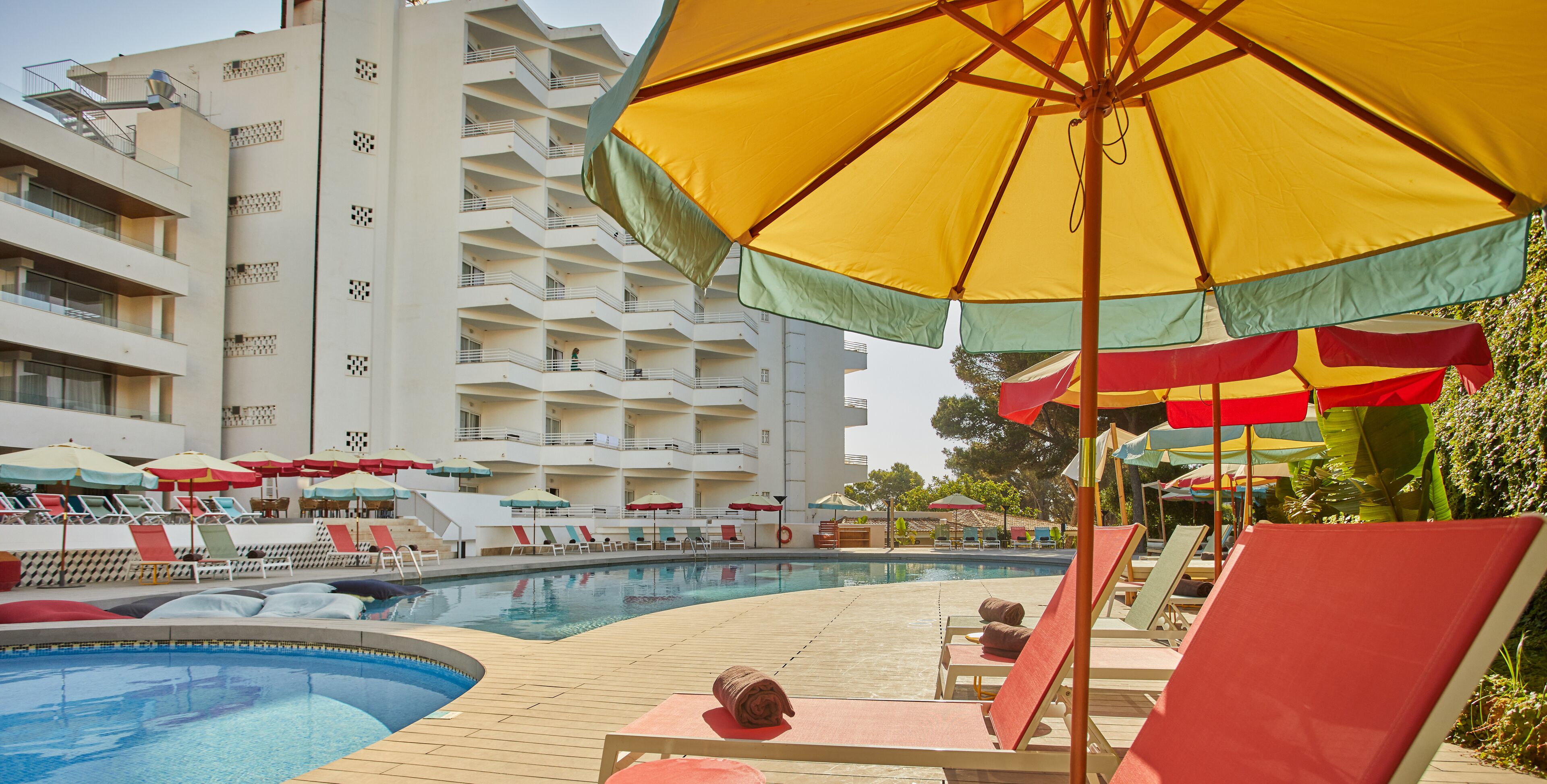 A hotel pool area with multiple lounge chairs and umbrellas, a large building with balconies in the background, and a clear sky.