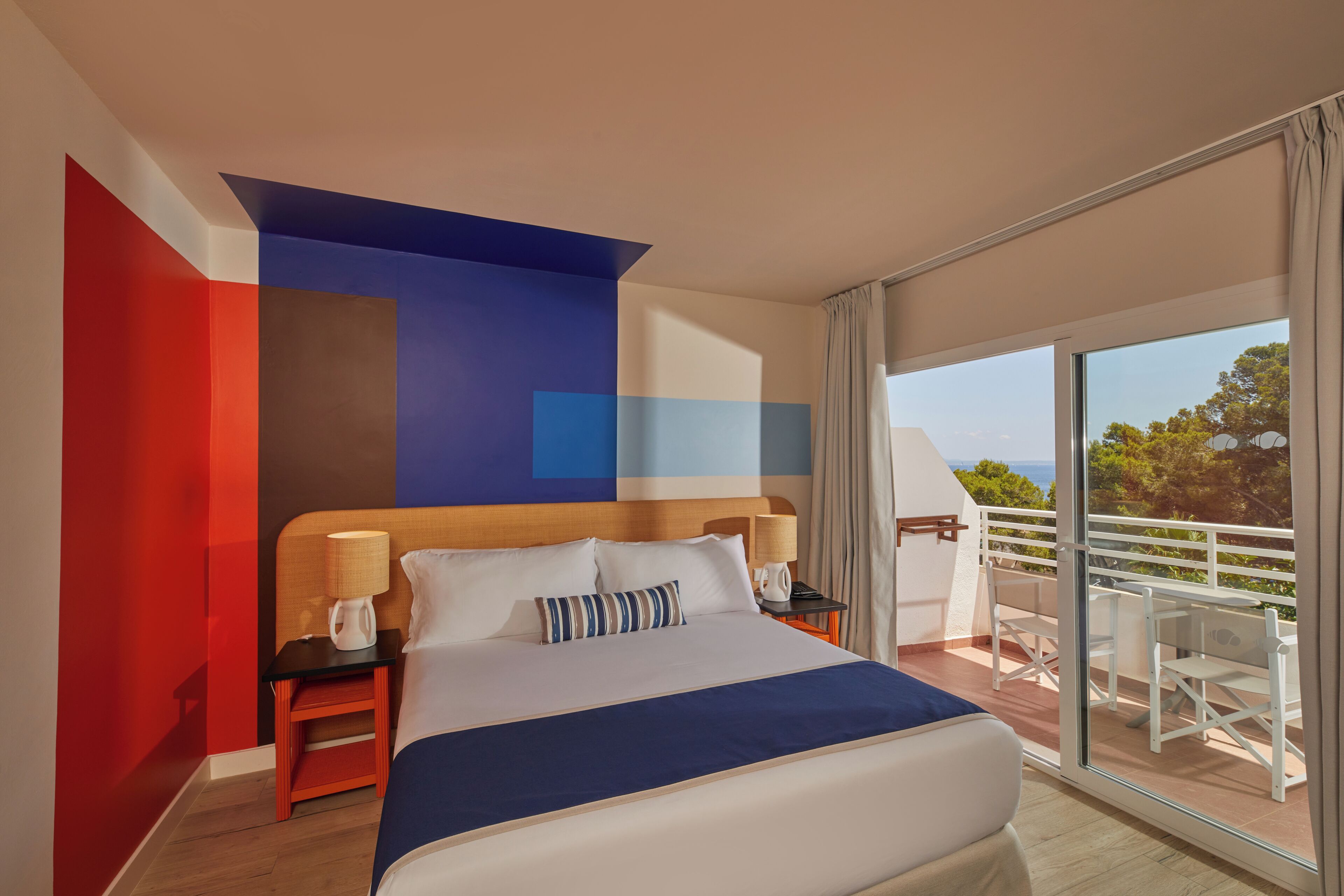 A modern hotel room with a large bed, a nightstand with a lamp, and a balcony with outdoor seating.