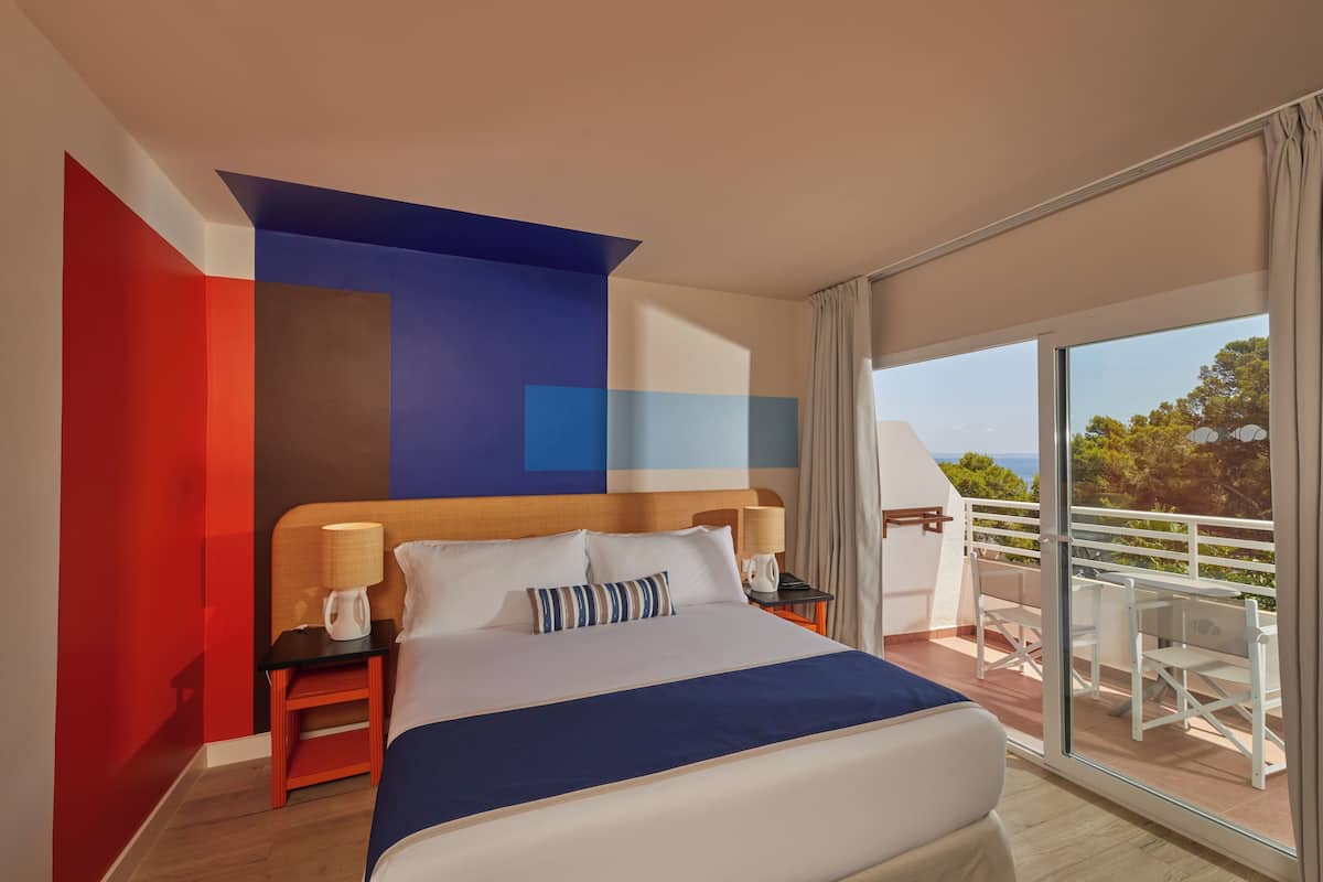 A modern hotel room with a large bed, a nightstand with a lamp, and a balcony with outdoor seating.