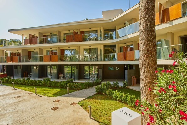 Un hotel moderno de varios pisos con un balcón de vidrio, un jardín cuidado y un letrero que dice 'PALM'.