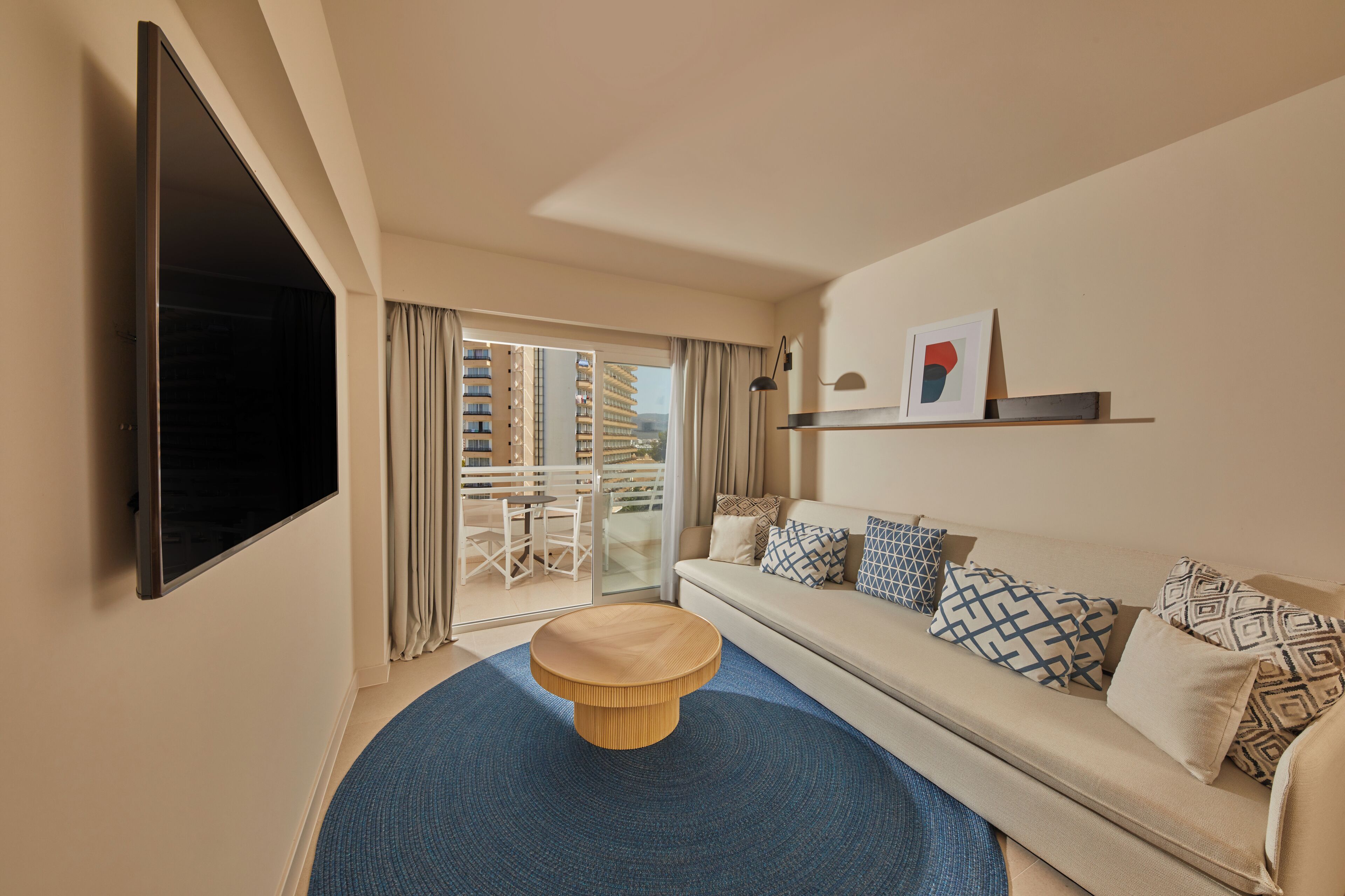 Un moderno cuarto de hotel con un televisor grande, un sofá con cojines estampados, una mesa de centro redonda de madera y vista a un edificio por la puerta de vidrio.