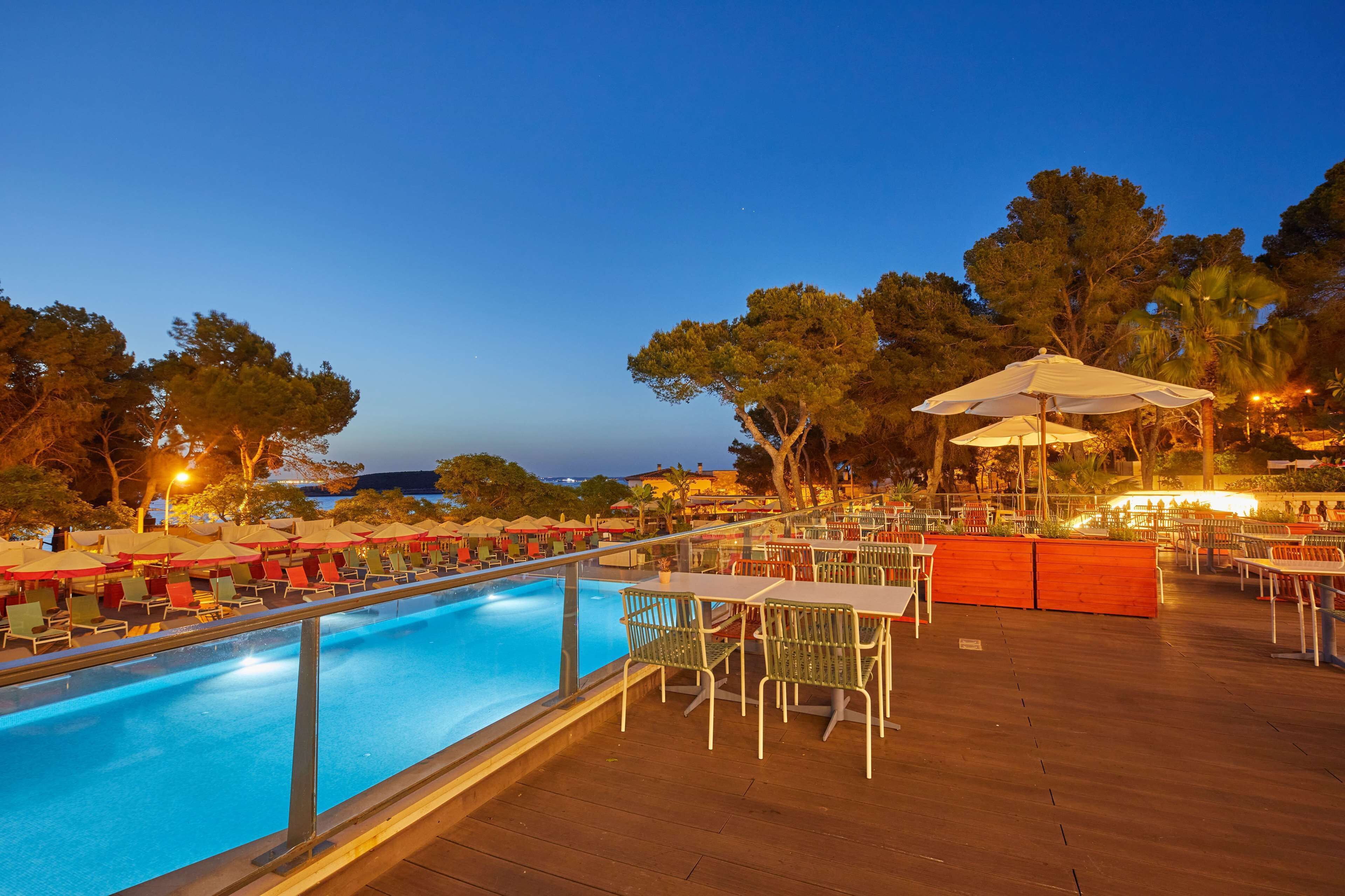 A poolside area with tables and chairs, a pool, and a view of the surrounding trees and buildings.