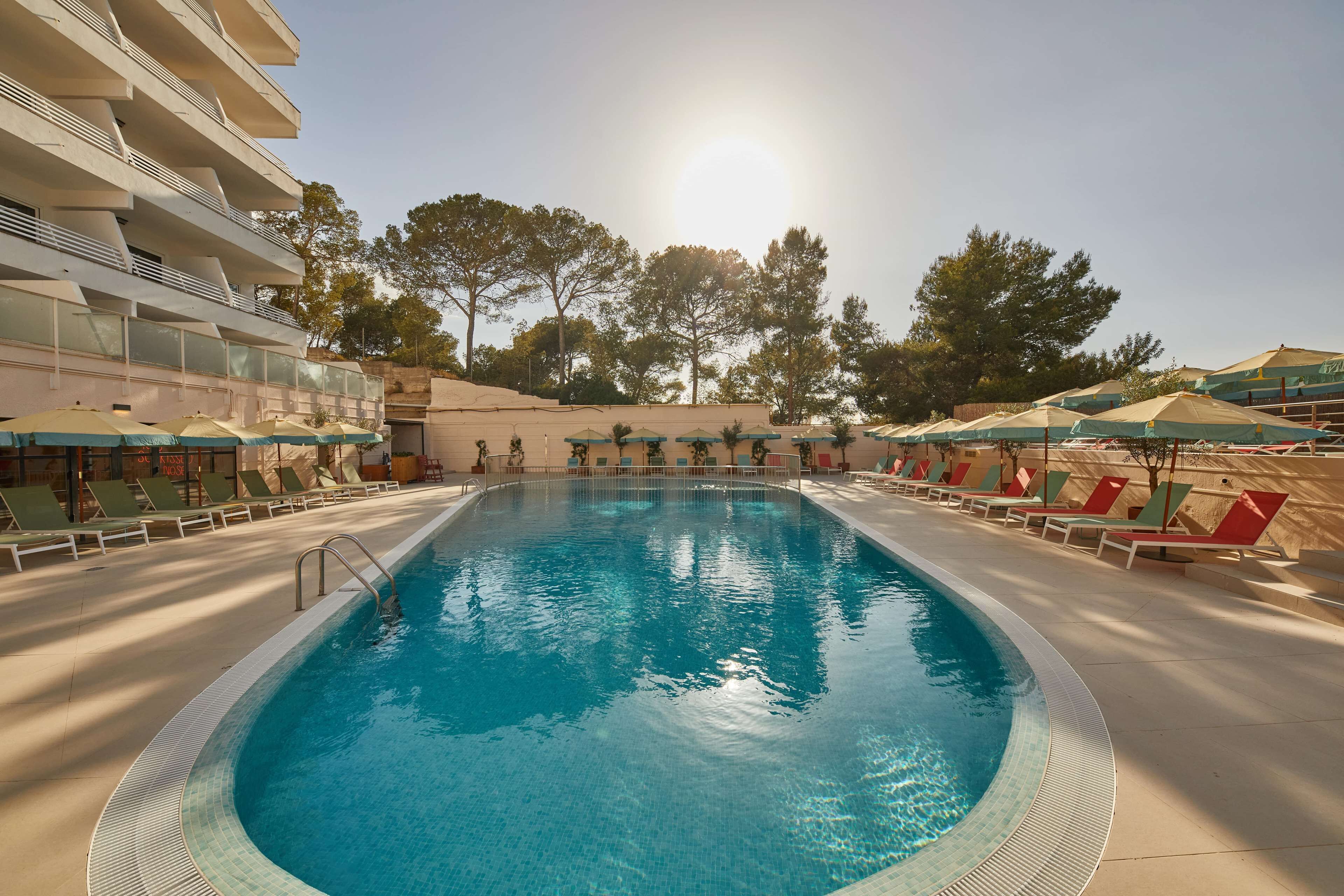 A swimming pool with sunlit water, surrounded by lounge chairs and umbrellas, and a building with balconies in the background.
