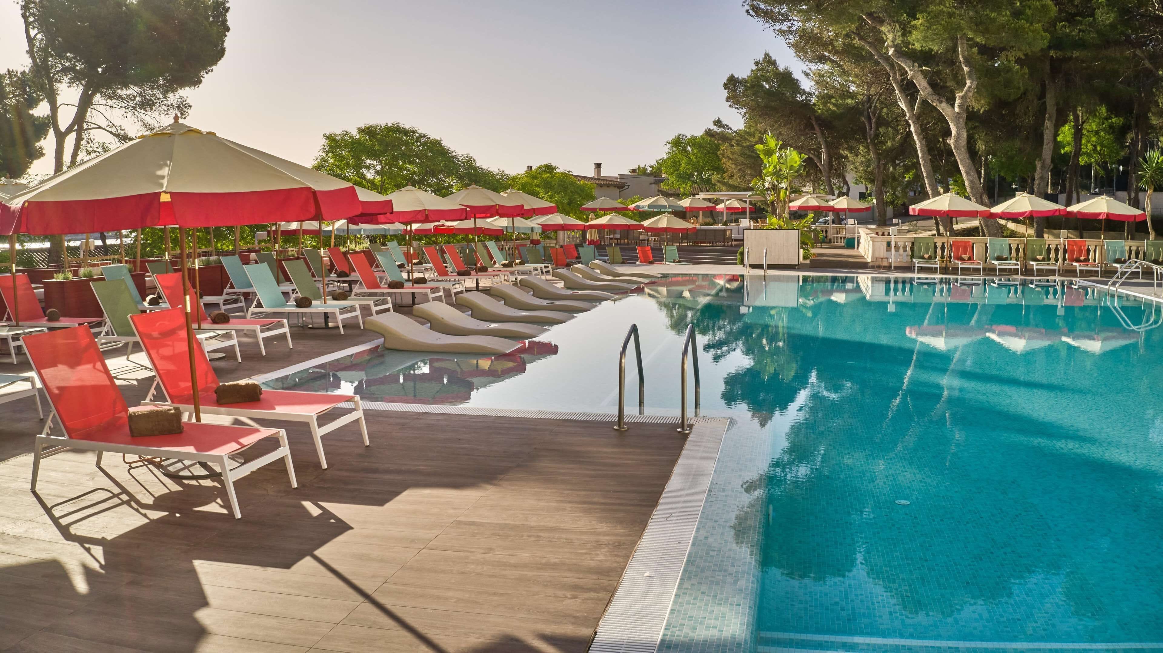 A poolside area with lounge chairs and umbrellas, a clear pool, and trees in the background.
