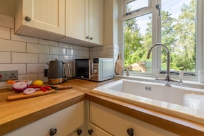 Une cuisine avec des meubles blancs, un plan de travail en bois, un évier, une fenêtre et une vue sur la verdure à l’extérieur.