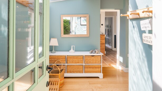 Un pasillo con pared azul claro, una consola blanca con cestos de mimbre, un espejo y una puerta que da a otra habitación.