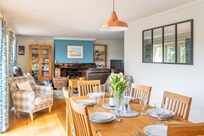 Un coin repas avec une table en bois dressée pour quatre personnes, entourée de chaises en bois, un vase rempli de fleurs et une cheminée en arrière-plan.