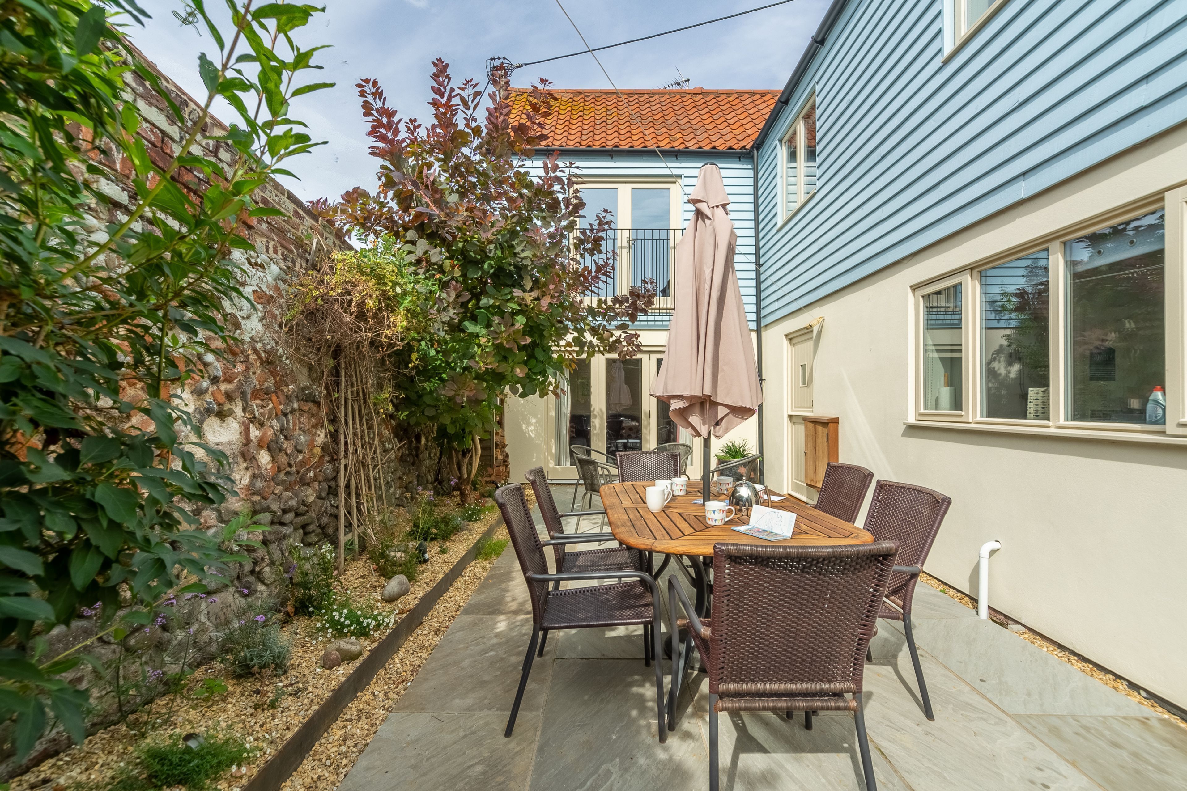A patio with a wooden table and wicker chairs, surrounded by a garden with plants and a stone wall.