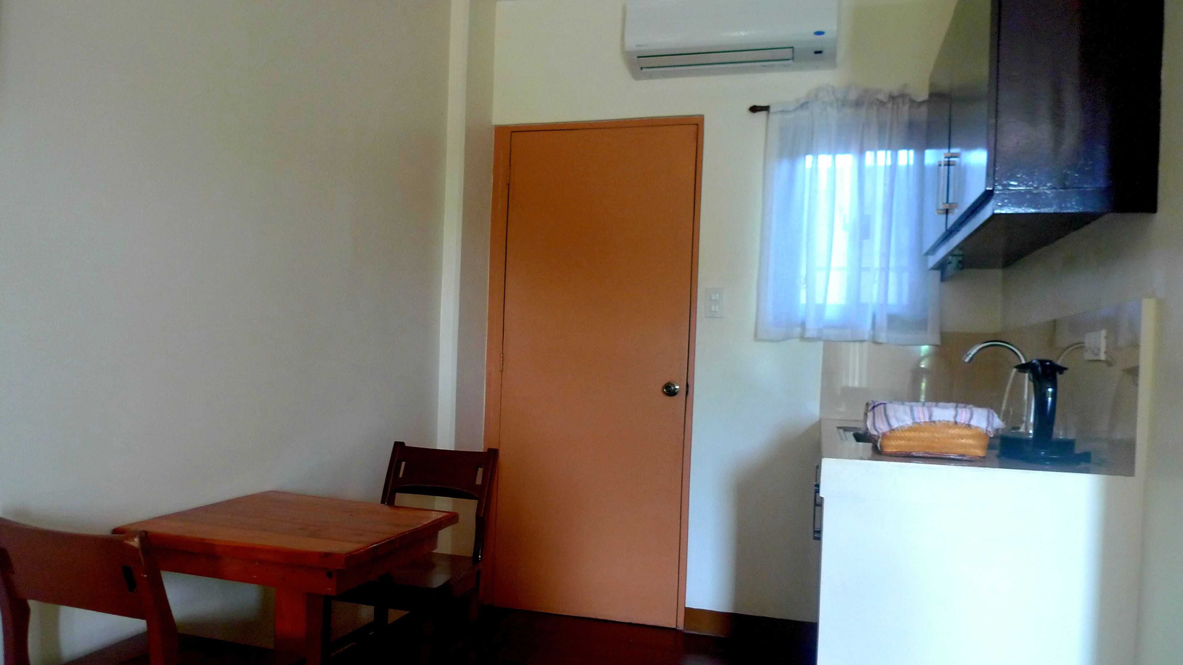 Une cuisine de taille modeste, avec une table et des chaises en bois, un réfrigérateur blanc et une fenêtre agrémentée d’un rideau.