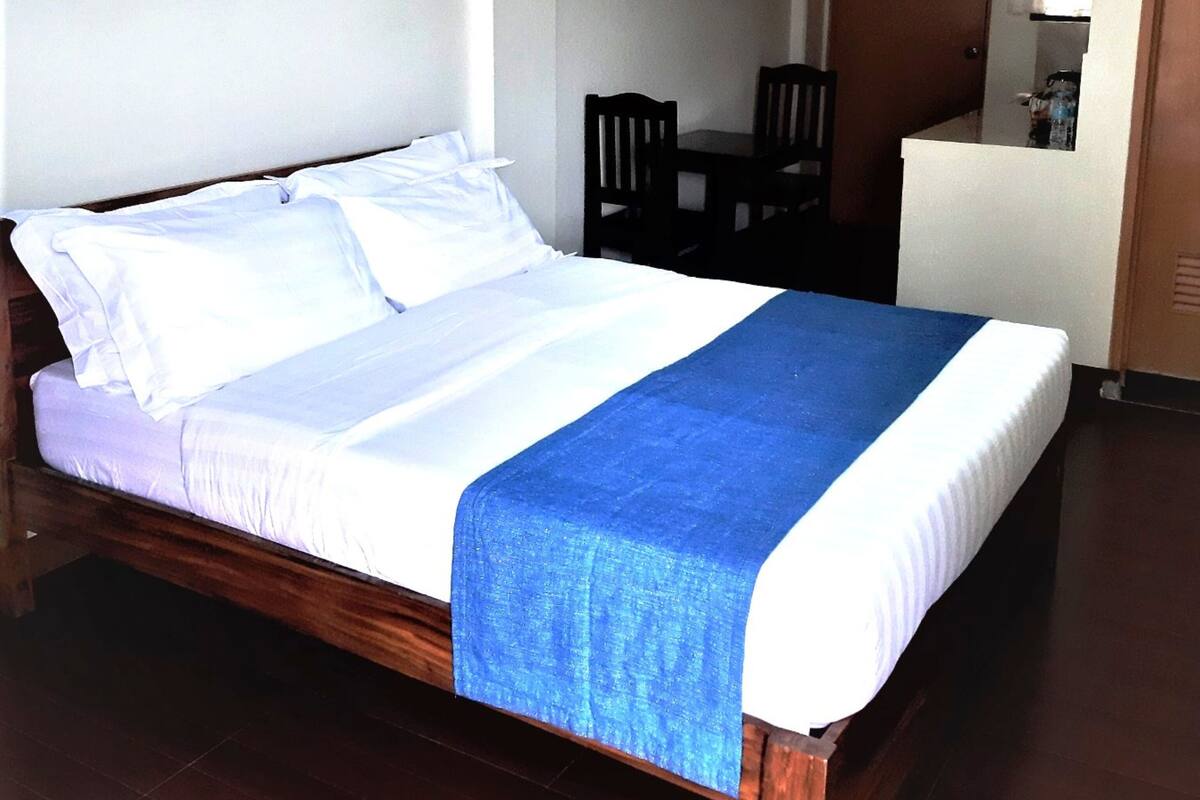 Ein Hotelzimmer mit einem hölzernen Bett, einem kleinen Tisch mit zwei Stühlen, einer weißen Wand und einer Tür zu einer Küchenzeile.